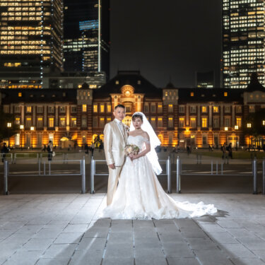 Wide shot of the couple standing on the plaza in front of the illuminated Tokyo Station, framed by glowing office towers, from a pre-wedding photoshoot in Tokyo.