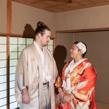 Groom in a cream montsuki and hakama smiling at his bride in a red crane uchikake inside a tatami room with shoji screens during their Japan wedding photography session