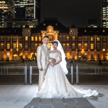 Full-length night portrait of the bride and groom with her long train spread on the paving stones, the warmly lit Tokyo Station building behind them, a classic Japan wedding photography composition.