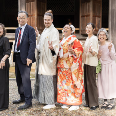 Couple in red uchikake and hakama laughing with their family in front of a historic wooden building, a joyful family shot from their kimono wedding photos in Japan