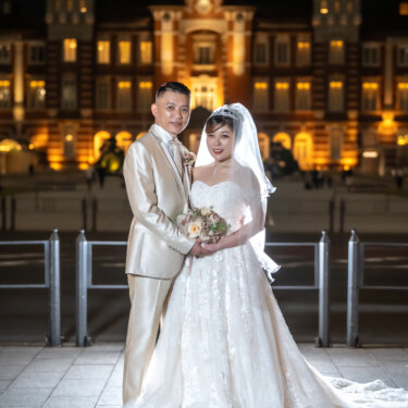 Couple in a champagne tuxedo and off-shoulder lace gown with a long veil standing close before the lit red-brick facade of Tokyo Station at night, a timeless wedding photo in Japan.