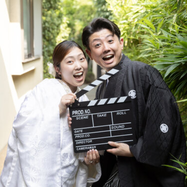 Bride in shiromuku and groom in montsuki laughing while holding a film clapperboard beside bamboo, a playful idea for a pre-wedding photoshoot in Japan