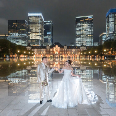 Bride and groom holding hands on a mirror-like water reflection with Tokyo Station and the Marunouchi skyscrapers glowing behind them in this night wedding photography shot.