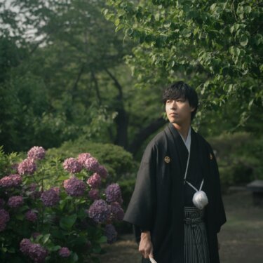 Groom in black montsuki and striped hakama holding a folding fan beside blooming hydrangeas on a garden path at Sankeien Garden, Yokohama