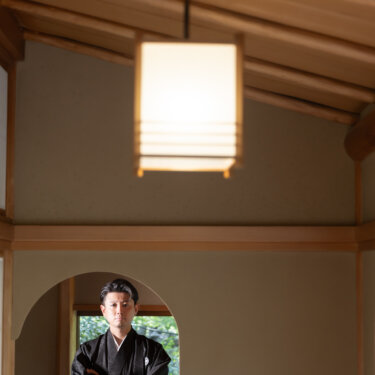 Groom in a black montsuki haori and striped hakama standing with arms folded beneath a glowing paper lantern, a dignified solo portrait from a Japanese wedding photoshoot