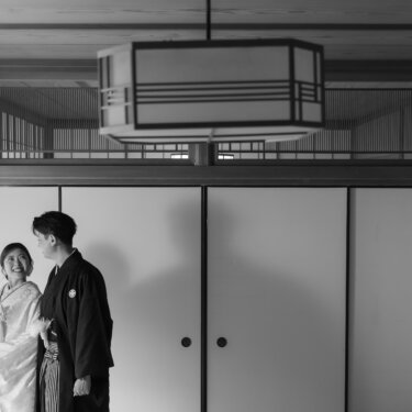 Black and white portrait of a couple in shiromuku and montsuki standing beside sliding fusuma doors under a modern Japanese lantern