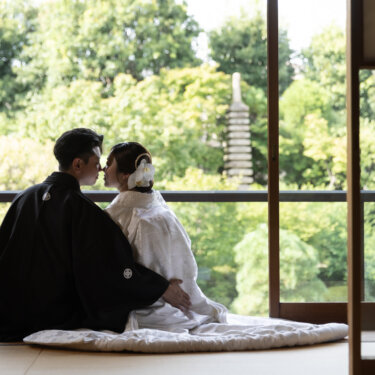 Bride and groom seated by open shoji doors with a stone pagoda and lush greenery behind them, about to share a kiss during their kimono photoshoot