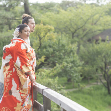 Couple in kimono gazing over the pond from a wooden bridge, with a thatched pavilion and early summer greenery behind them in this Japan wedding photography shot