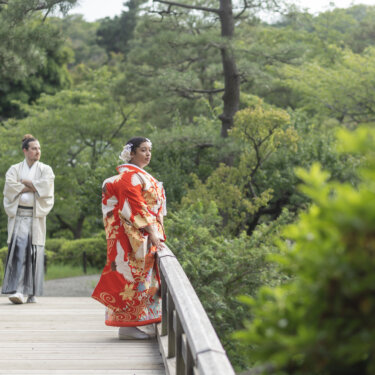 Bride leaning on the bridge railing in a red crane uchikake while the groom in hakama walks toward her, a relaxed moment from a kimono wedding photoshoot at Sankeien Garden