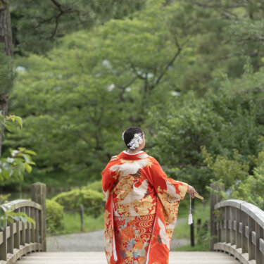 Back view of the bride in a vivid red uchikake with white flower kanzashi, spreading her sleeves on a wooden bridge in a Japanese garden for her wedding photos