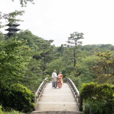 Couple in kimono standing on a long wooden bridge with the three-story pagoda rising above the pine trees, a wide shot from their pre-wedding photoshoot in Japan