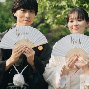 Bride in shiromuku and groom in black montsuki holding white folding fans that read 'Groom' and 'Bride' in a green Japanese garden in Yokohama