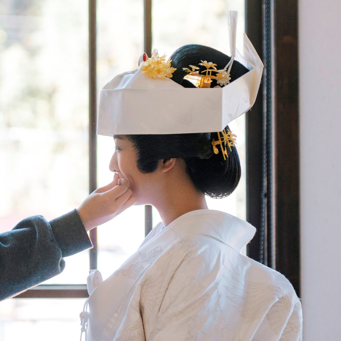 The bride, dressed in a pure white shiromuku with a tsunokakushi headdress, smiles as an attendant adjusts her makeup by a window.