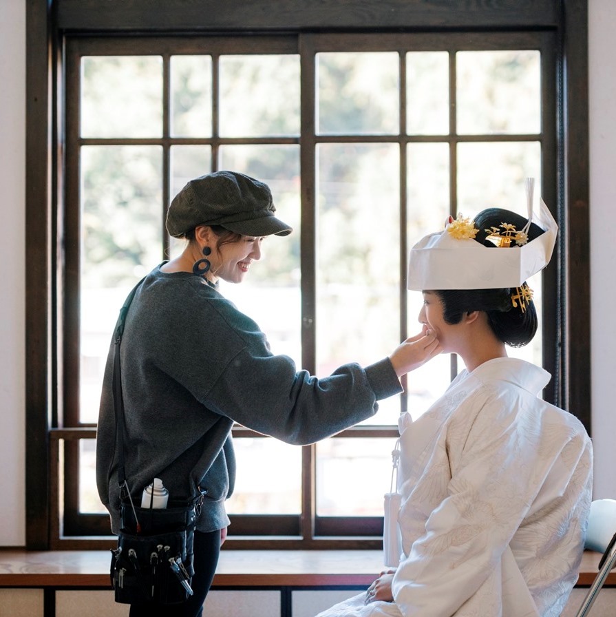 Hairstylist Nanako adjusts the bride's makeup by a large window, the bride in a white shiromuku and paper headdress.