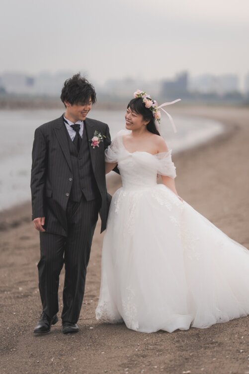 Smiling couple strolling along the beach, the groom in a black pinstripe tuxedo and the bride in a flower crown, captured as wedding photos in Japan (TP08).