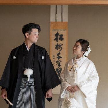 Bride in a white shiromuku with a silk bridal purse and groom in striped hakama exchanging glances in a tatami room captured in this pre-wedding photoshoot in Japan