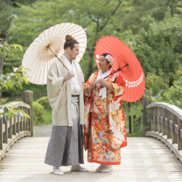 Bride in a red crane-patterned uchikake and groom in a cream hakama holding red and white wagasa umbrellas on a wooden arched bridge at Sankeien Garden, a kimono wedding photos highlight in Yokohama