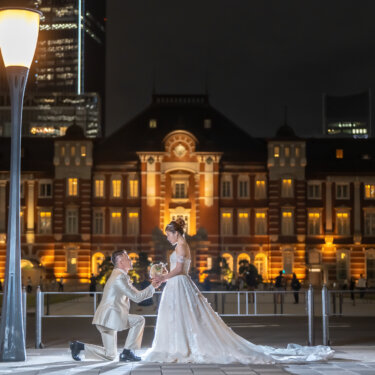 Groom in an ivory tuxedo kneeling to offer his bride a bouquet in front of the illuminated Tokyo Station Marunouchi building, a romantic proposal-style pose for a night pre-wedding photoshoot in Japan.