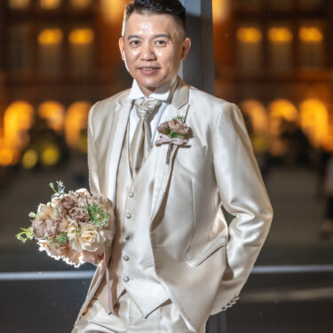 Portrait of the groom in a champagne three-piece tuxedo holding the bouquet with one hand in his pocket, the golden lights of Tokyo Station glowing behind him during a night wedding photoshoot.