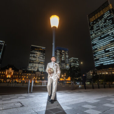 Groom in an ivory tuxedo walking beneath a street lamp on the Marunouchi plaza at night, with Tokyo Station and the high-rise towers lit up behind him, from a pre-wedding photoshoot in Japan.