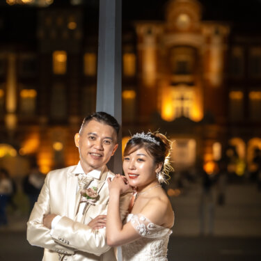 Bride hugging her groom from the side with a smile against the softly blurred lights of Tokyo Station at night, a warm couple portrait from their wedding photoshoot.
