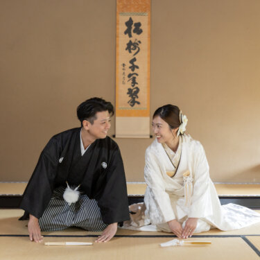 Bride and groom kneeling on tatami and laughing together in front of a hanging scroll, relaxed moment from a traditional Japanese wedding photoshoot