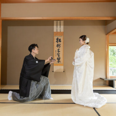 Groom in black montsuki kneeling on tatami to offer a ring box to his bride in a white shiromuku, a proposal scene staged in a Japanese tatami room