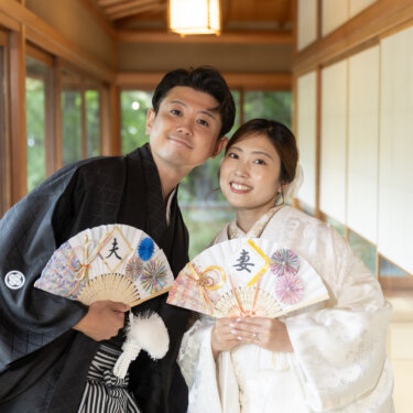 Couple in traditional wedding kimono holding decorative fans and smiling in a wooden corridor overlooking a garden in this kimono wedding photos in Japan