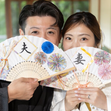 Bride and groom peeking over paper fans painted with the characters for husband and wife, a playful close-up from a traditional Japanese pre-wedding photoshoot