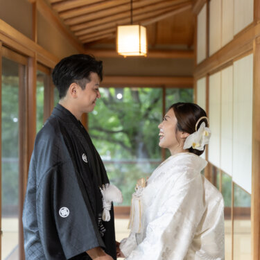 Smiling couple holding hands in a sunlit wooden corridor, the bride in a shiromuku with an orchid hair ornament and the groom in a black montsuki haori hakama during their Japan wedding photography session