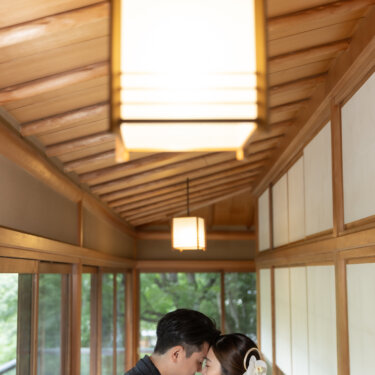 Bride in a white shiromuku kimono and groom in black montsuki touching foreheads beneath a wooden lantern in a traditional Japanese corridor, a quiet moment from a kimono wedding photoshoot in Japan