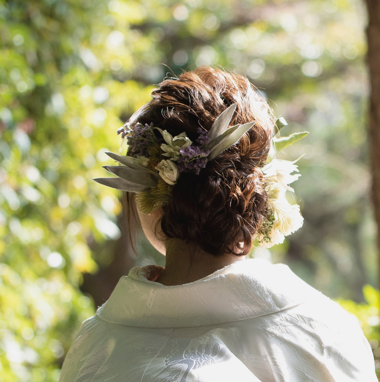 A romantic bridal updo woven with lavender statice and cream dried flowers against a sunlit garden backdrop.