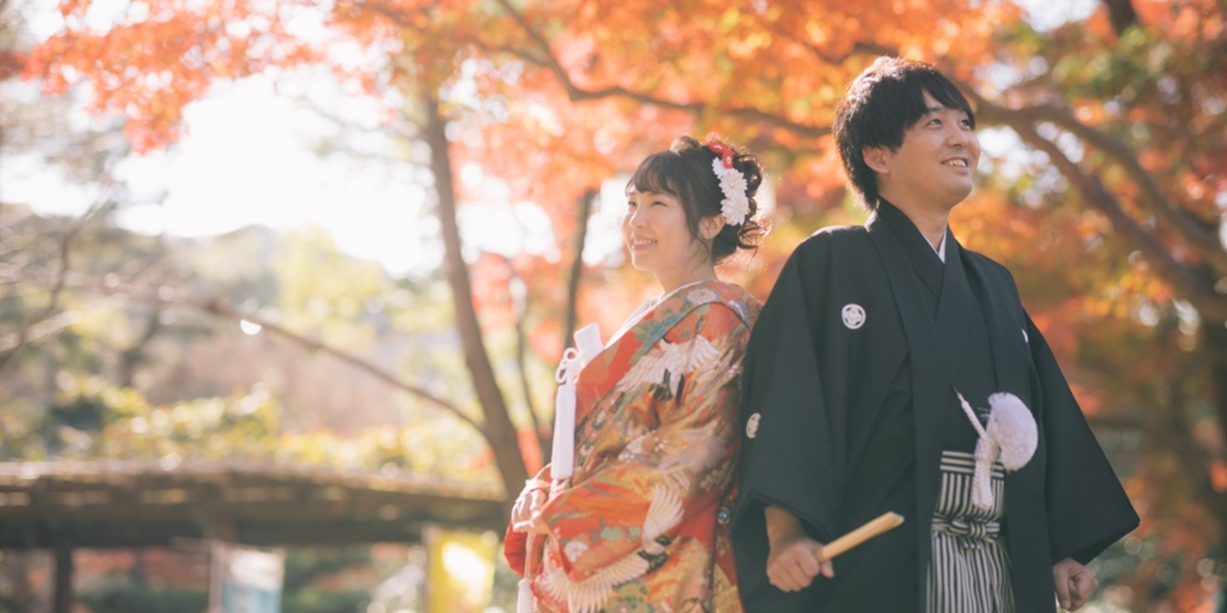 Standing back to back beneath fiery autumn maples at Gifu Park, the bride's vivid crane-patterned kimono glowing in the light during this Japan wedding photography session.