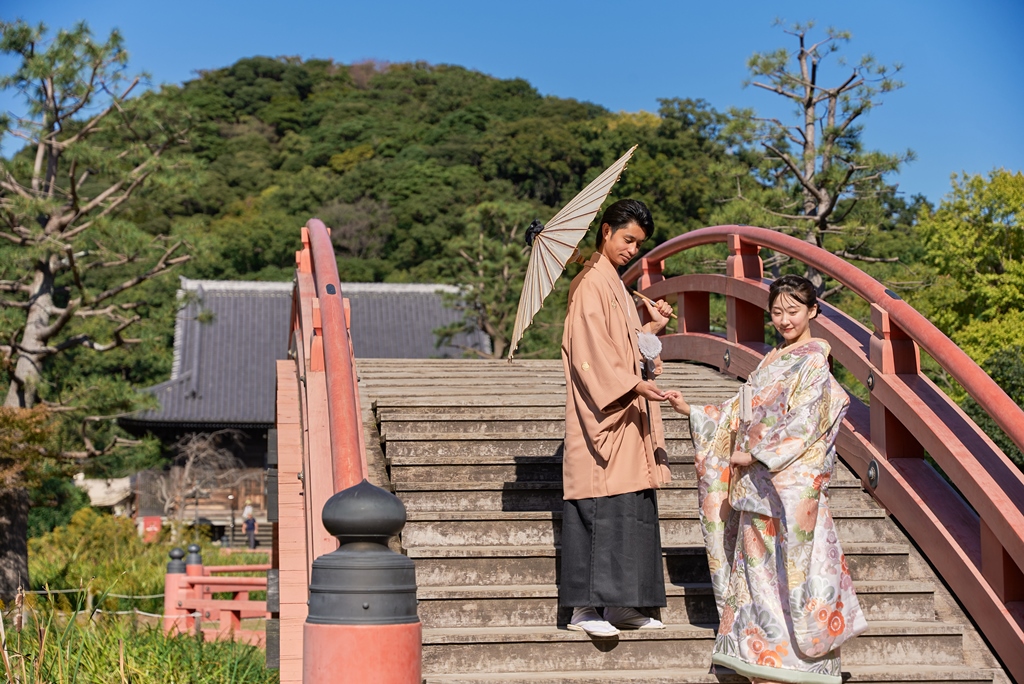 The couple stands on a red arched bridge, the groom holding a paper umbrella over the kimono bride.