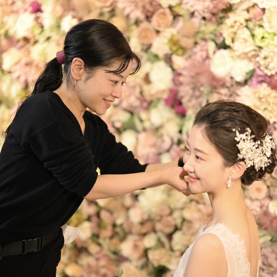 Hair and makeup artist Tada gently touches up the bride's face, a wall of pink and cream flowers behind them.