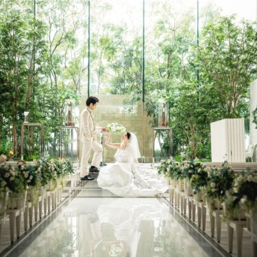 Bride in a ball gown seated on a mirrored aisle as the groom takes her hand in a glass chapel filled with greenery, with their reflection below