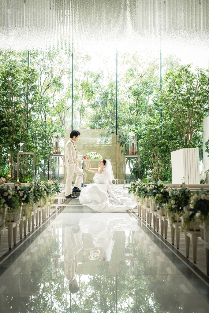 Bride in a ball gown seated on a mirrored aisle as the groom takes her hand in a glass chapel filled with greenery, with their reflection below