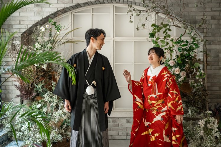 Couple laughing together before a white brick arch framed with flowers, the groom in montsuki hakama and the bride in a red crane uchikake — Japan wedding photography