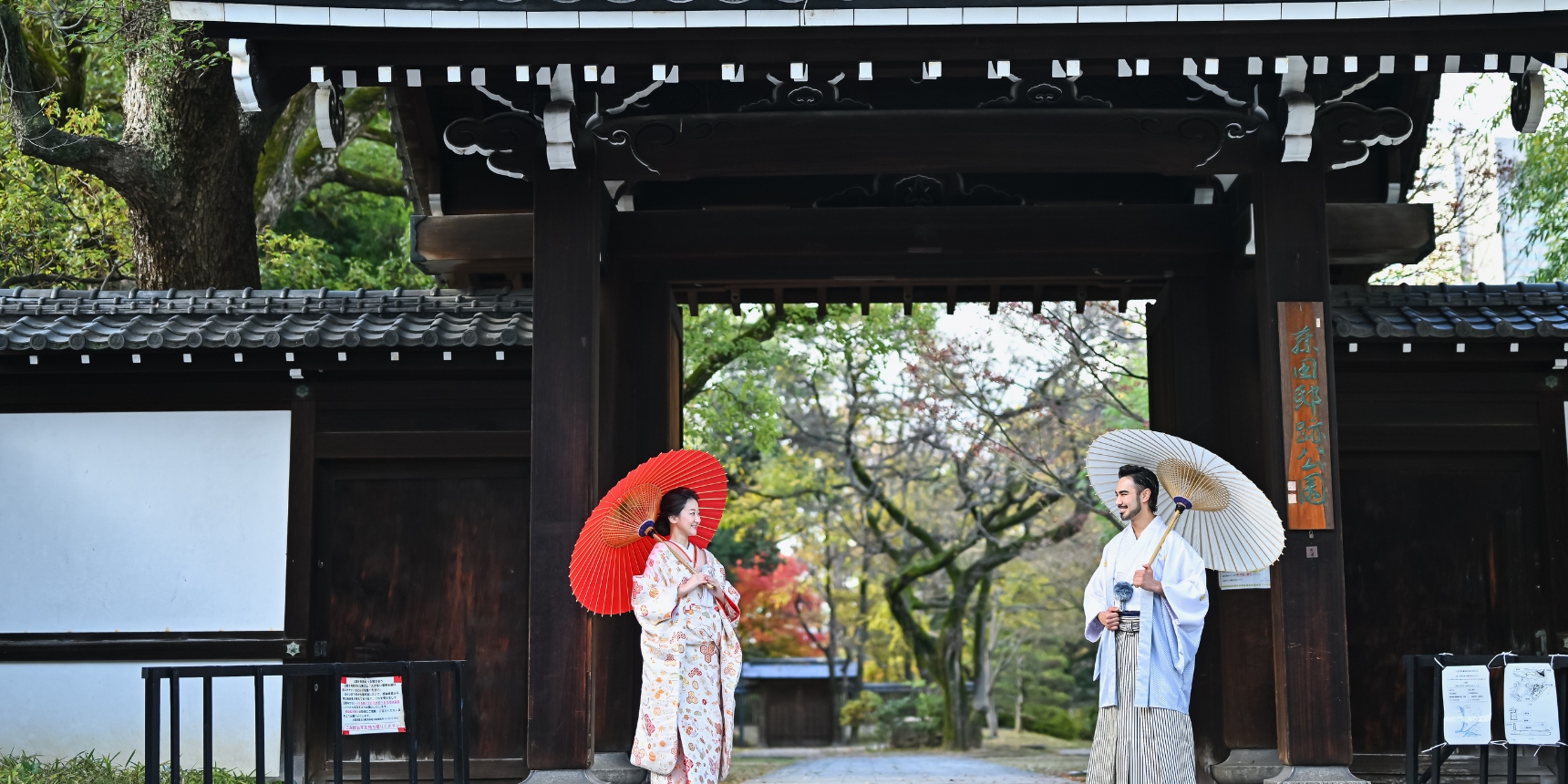 Holding red and white paper umbrellas beneath the towering wooden gate of Fujita-tei Ato Park, autumn maples glowing in the distance.