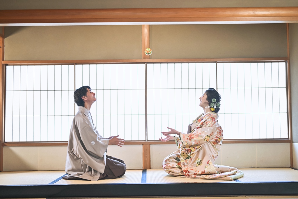 The bride and groom toss ball-shaped charms toward each other, laughing in a tatami room.
