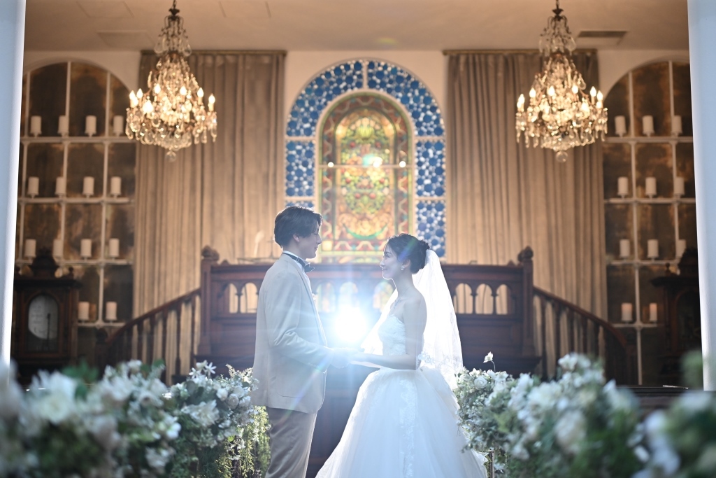 Couple standing hand in hand at the altar of a grand chapel, lit by chandeliers and a stained-glass window, white floral aisle decorations in the foreground.