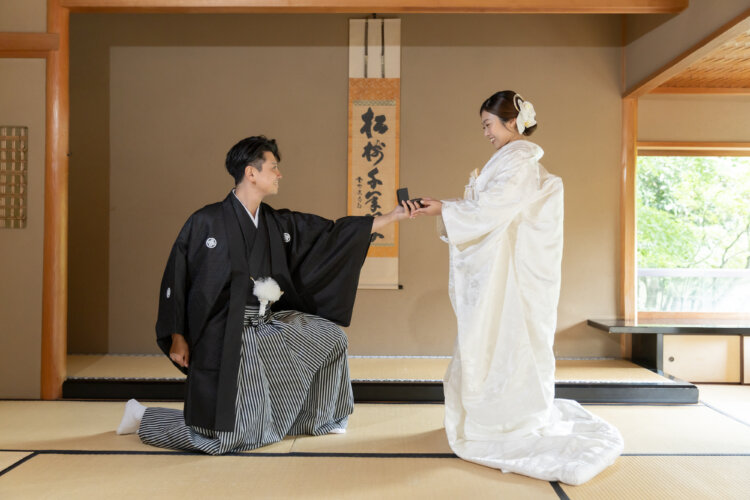 Groom in black montsuki kneeling on tatami to present a ring box to his bride in shiromuku, a proposal scene recreated in a traditional Japanese room