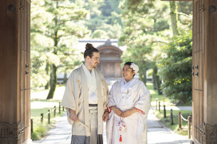 Smiling bride in shiromuku and groom in hakama standing together in an open wooden gateway with the garden path behind them, a welcoming frame for kimono wedding photos