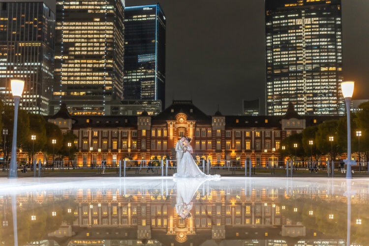 Tiny silhouette of the bride and groom on a glowing water mirror before the illuminated Tokyo Station, the reflection doubling the night scene in this Japan wedding photography shot.