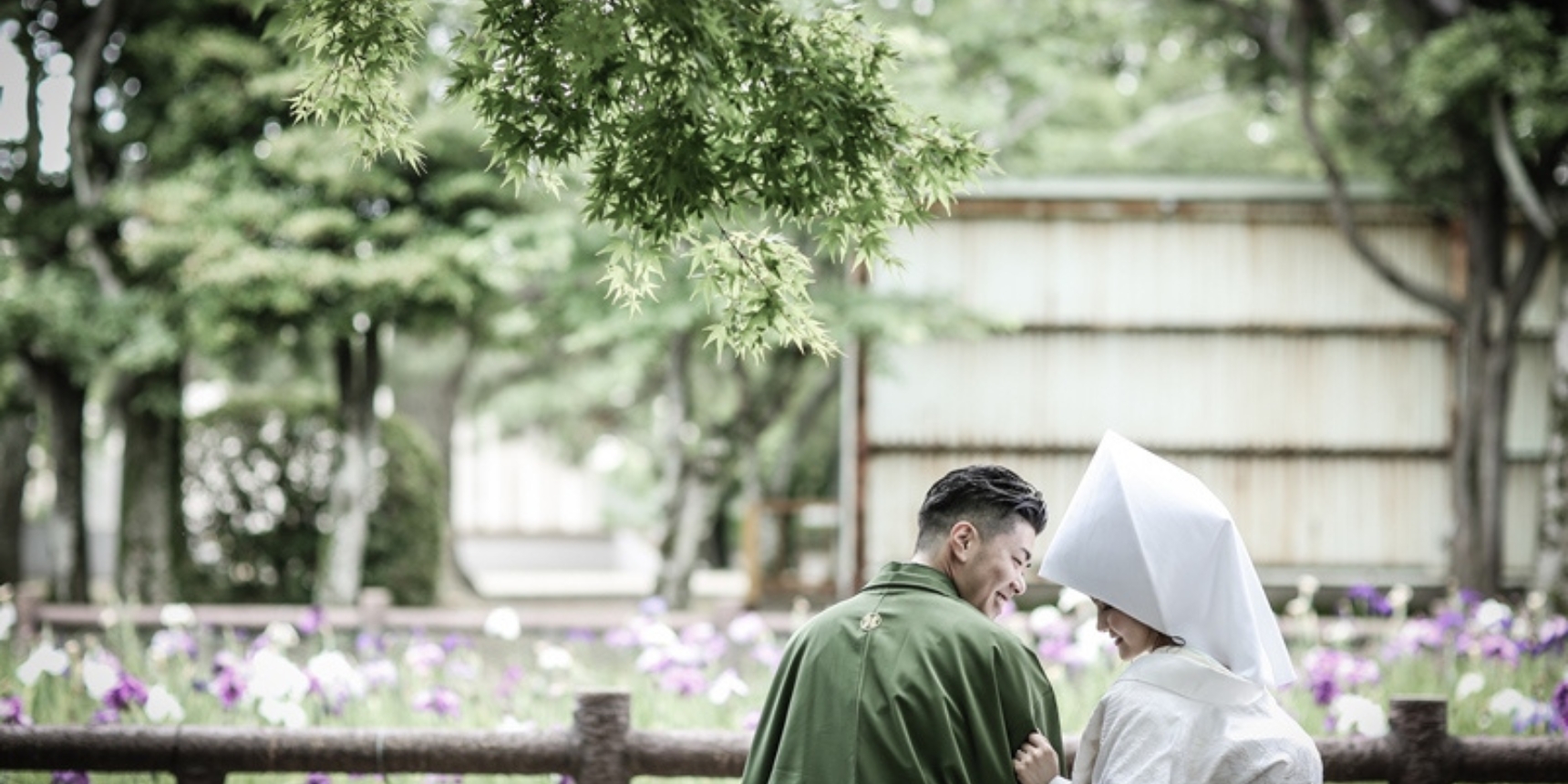 Walking together past blooming purple and white irises at Kyuka Park, the bride wearing a traditional white tsunokakushi headdress.