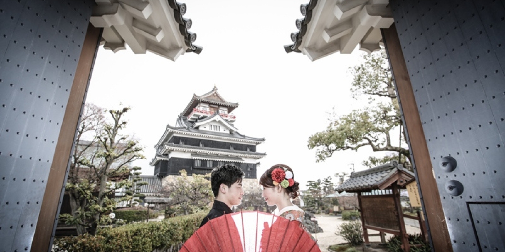 Framed by the gate of Kiyosu Castle, the bride holds a bold red fan as the historic keep rises behind them, a striking scene for wedding photos in Japan.