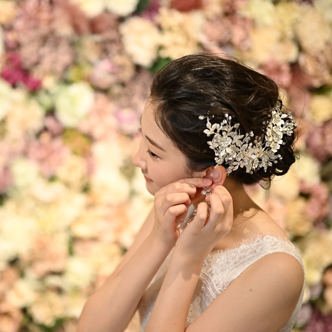 A bride adjusts her earring, wearing a sparkling floral hair accessory against a flower wall backdrop.