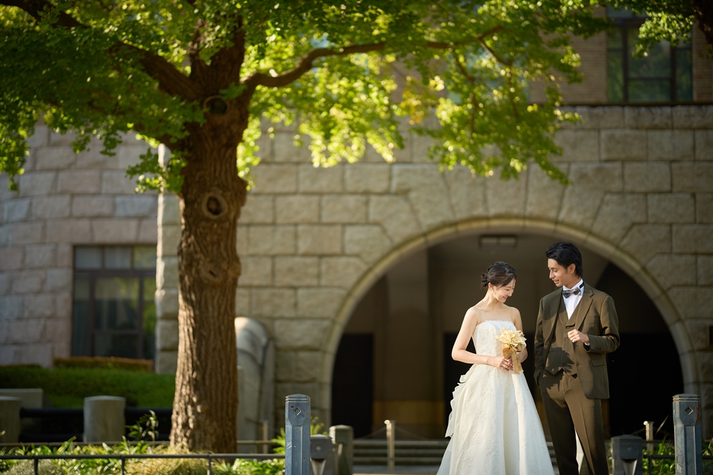 The bride and groom stroll past a stone archway building, a tall tree shading the sunlit street.