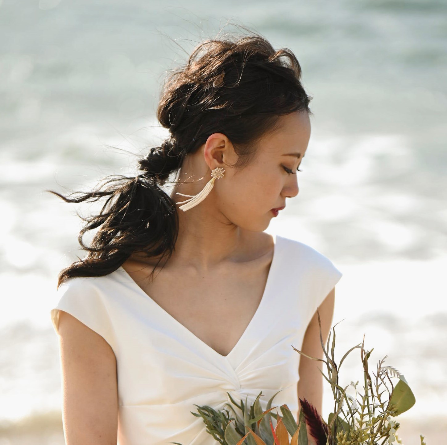 The bride's wind-swept ponytail updo catches the ocean breeze, paired with delicate tassel earrings and an off-shoulder ivory gown.
