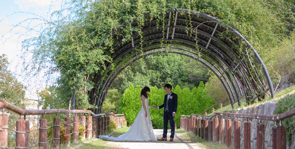 Walking hand in hand beneath a vine-covered arch tunnel at Moricoro Park, greenery framing the path ahead.